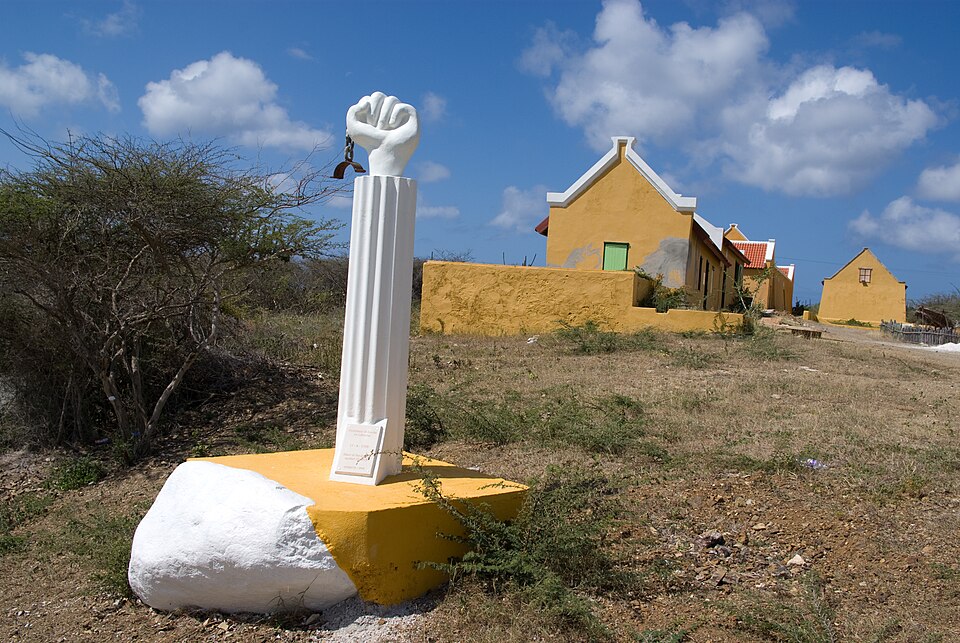 Monumento blanco con un puño en alto a la rebelión de 1795, frente a la casa amarilla de Landhuis Kenepa