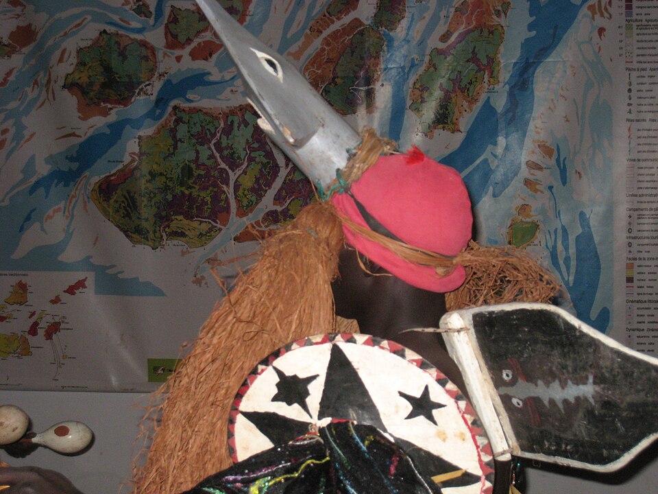Máscara tradicional bijagó con cuernos y escudo pintado frente a un mapa del archipiélago