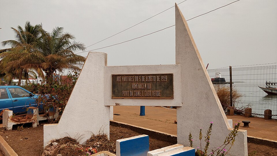 Monumento blanco con una placa en memoria de la masacre de Pidjiguiti junto al puerto