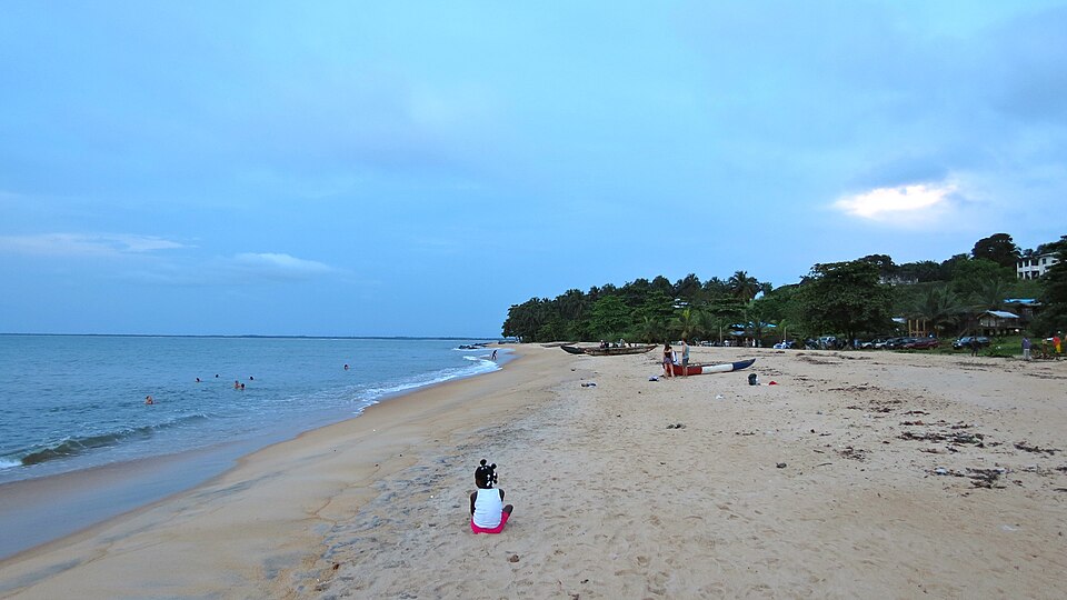 Playa larga de arena en Robertsport con bañistas y árboles al fondo