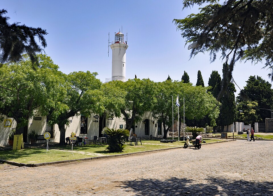 Plaza arbolada del barrio histórico de Colonia del Sacramento con el faro blanco al fondo